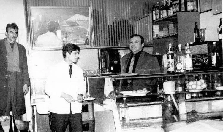 Crno-bela fotografija: konobar u belom sakou i gosti kod šanka sa vitrinom