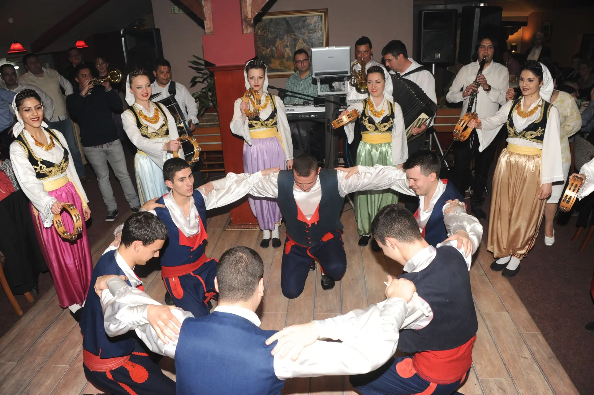 Igrači folklora kleče u krugu, iza njih igračice sa defovima i orkestar